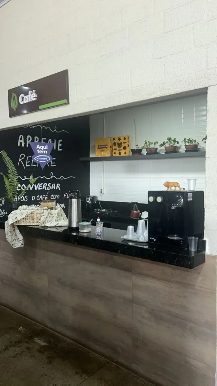 O cantinho do café da loja: lousa preta escrita à mão, garrafa térmica, máquina de café e vasinhos de planta sobre o balcão de granito